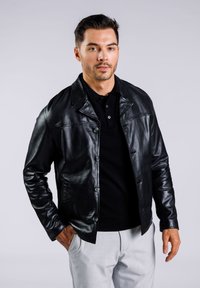 Svart läderjacka med knapplåsning fram, vikkrage och skräddarsydd passform. Bärs över en svart polo, tillsammans med ljusgrå byxor.