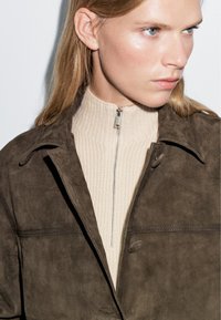 Giacca in suede marrone con colletto a lobo, abbinata a un maglione beige a coste con zip. Caratterizzata da zip eleganti e una texture liscia.