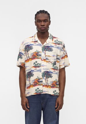 Man with braided hair wearing a short-sleeve button-up shirt with tropical palm tree and island landscape print and dark blue jeans.