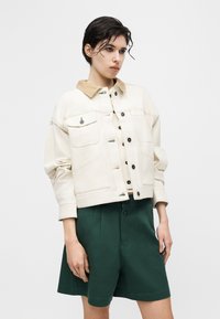 Νεαρό άτομο με κοντά σκούρα μαλλιά, που φοράει μια μπεζ ζακέτα με κουμπιά και καφέ γιακά, καθώς και σκούρα πράσινα σορτς μέχρι το γόνατο, στέκεται μπροστά σε λευκό φόντο.