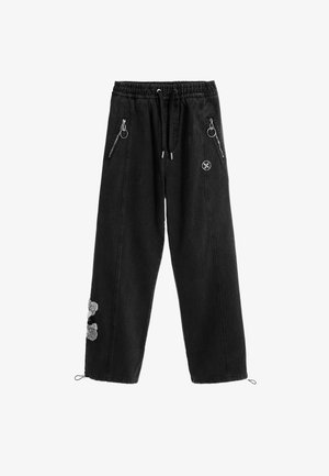 Pantalones de mezclilla negra con cintura elástica, cordón y bolsillos laterales con cremallera. Presenta un detalle de aplique peludo en una pierna.