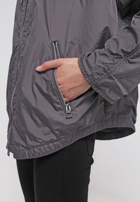 Veste légère grise avec une poche zippée, une texture lisse et une coupe décontractée. Comprend des poignets élastiques et un ourlet incurvé.