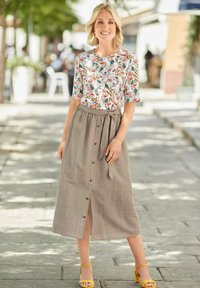 Blumenbluse mit kurzen Ärmeln, kombiniert mit einem beigen Leinenrock mit Knöpfen und einem Bindeband in der Taille. Getragen mit gelben Absatzsandalen.