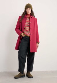 Fuksiasta volnen plašč z gumbom spredaj in širokimi revnimi, kombiniran s potiskanim puloverjem in črnimi visokim pasom cargo hlačami. Olivno zeleni škornji.