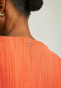Abito plissettato di colore corallo con un accento argento rotondo all'apertura sul retro, che mostra una finitura liscia e testurizzata.
