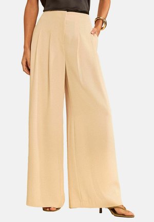 Beežid kõrge vöökohaga laia säärega püksid musta topi, avatud varvastega kontsakingade ja kuldse käevõruga, üks käsi vabalt taskus.