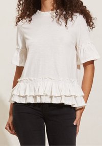 Bílá halenka s krátkým rukávem a volánkovým lemem a rukávy. Má texturované detaily podél výstřihu a okrajů rukávů. Lehká tkanina.