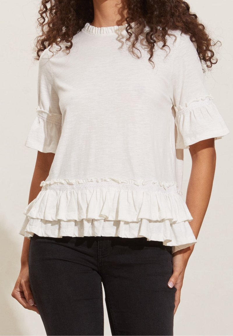 Bílá halenka s krátkým rukávem a volánkovým lemem a rukávy. Má texturované detaily podél výstřihu a okrajů rukávů. Lehká tkanina.