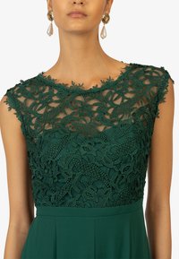 Emerald green dress featuring intricate lace detailing at the neckline, with a smooth skirt below. Accented with pearl drop earrings.