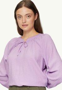 Lavendelblouse, mille tekstuuri, ümmarguse kaelusega ja lipsukinnitusega. Omab rüüstatud varrukaid ja lõdvemat istuvust.