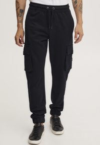 Zwarte katoenen cargo joggers met elastische boorden, een trekkoord in de taille, zijzakken en gestikte details. Gemaakt in combinatie met zwarte sneakers.
