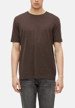 Mann trägt ein schlichtes dunkelbraunes kurzärmeliges T-Shirt und ausgewaschene schwarze Jeans, steht vor weißem Hintergrund, Arme entspannt an den Seiten.