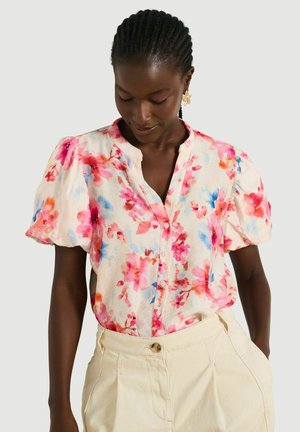 Vrouw met gevlochten haar die een korte mouw bloemenblouse draagt in roze en blauwe tinten, gecombineerd met crèmekleurige hoge broeken, kijkt naar beneden.