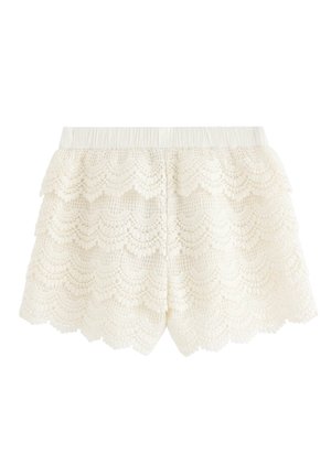 Ung pige stående iført brun flæsebluse, hvide blonde-shorts, cremefarvet cardigan og mørke sko med nitter foran en ensfarvet baggrund.