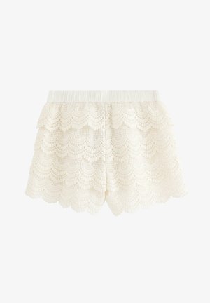 Młoda dziewczyna stoi, ubrana w brązowy top z falbankami, białe koronkowe szorty, kremowy kardigan oraz ciemne baleriny z ćwiekami na jednolitym tle.