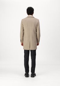 Hellbrauner Wollmantel mit taillierter Passform, langen Ärmeln und Reverskragen. Kombiniert mit schwarzen Hosen und schwarzen Schuhen.