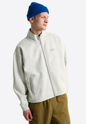 Giovane uomo che indossa un berretto di lana blu, una giacca grigio chiaro con zip e tasche e pantaloni marroni, guarda di lato con un leggero sorriso.