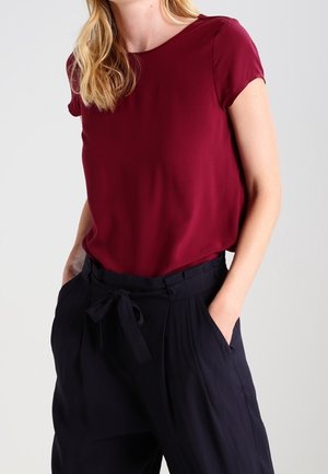 Kobieta w bordowej bluzce z krótkim rękawem, wsuniętej w wysokiej talii czarne spodnie z paskiem wiązanym, ręce w kieszeniach, kadrowana na wysokości ramion i bioder.
