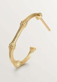 Pendiente de aro en tono dorado, que recuerda a una estructura ósea. Presenta un acabado texturizado, con detalles estriados y un diseño abierto con un pasador recto.