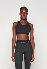Top desportivo preto com alças largas e textura suave, combinado com leggings escuras com padrão, apresentando uma cintura preta com o logótipo "new balance".
