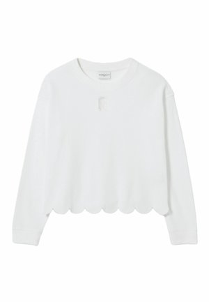 Femme portant un sweat blanc à ourlet festonné, short rose clair, baskets rouges, tenant un sac à main noir, debout sur un fond uni.