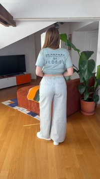 Oseba stoji v notranjosti z obrazom stran, oblečena v svetlo modro kratko majico in svetlo sive široke hlače, blizu velika zelena sobna rastlina v lončku.