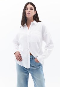 Weißes Button-Up-Hemd aus leichtem Stoff mit Brusttasche, langen Ärmeln und bequemer Passform, kombiniert mit hellblauen Jeans.