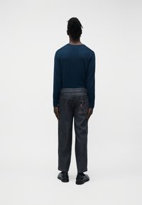 Personne debout, dos à la caméra, portant une chemise à manches longues bleu marine, un jean foncé avec une broderie d'aigle sur la poche arrière droite, et des chaussures noires.