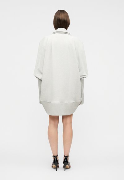 Γυναίκα με πλάτη γυρισμένη, φοράει ελαφρύ oversized φόρεμα πουλόβερ και μαύρα πέδιλα με ψηλό τακούνι σε απλό φόντο.