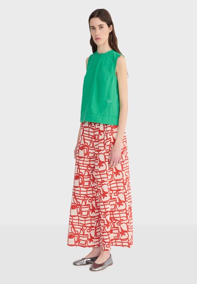 Femme debout portant un haut vert sans manches, un pantalon blanc à jambes larges avec un motif abstrait rouge, et des chaussures plates argentées sur un fond uni.