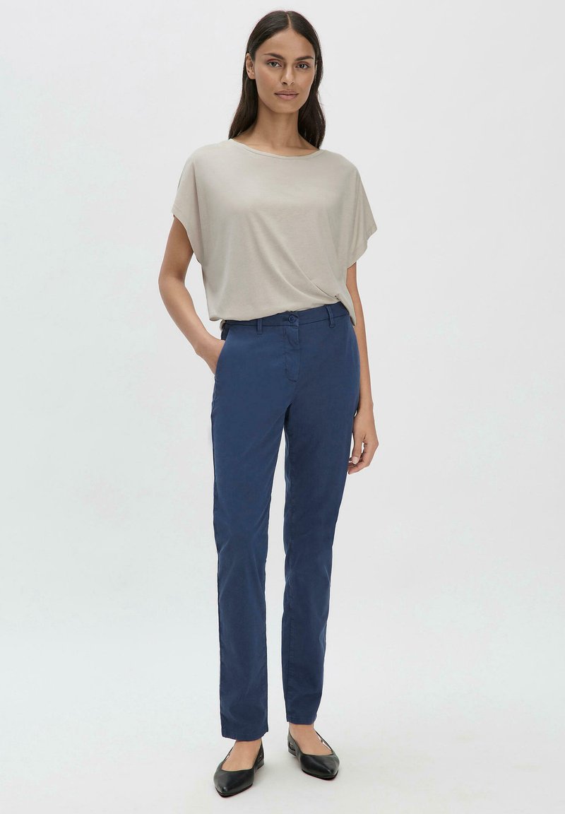 Lichtbeige top met een losse pasvorm en wijde mouwen, gecombineerd met slimfit marineblauwe broeken. De outfit heeft minimale details en klassieke zwarte flats.