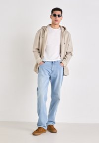 Beige zipzáras kapucnis pulóver, fehér póló, világoskék farmernadrág, barna tornacipő és fekete napszemüveg. A modell egy sima háttér előtt áll.