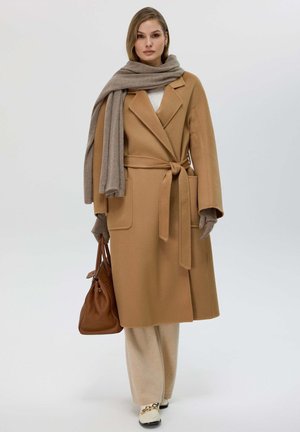 Manteau en laine couleur camel avec une taille à lien, associé à une écharpe beige et un pantalon assorti. Il possède de grandes poches et une texture lisse.