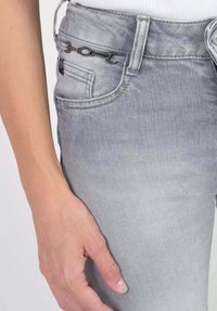 Jean en denim gris avec un effet délavé. Comprend une poche latérale, un bouton en métal et une chaîne décorative à la taille. Texture lisse.