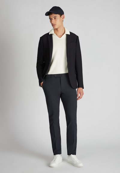 Giovane uomo che indossa un blazer nero, un polo in maglia bianca, pantaloni neri, sneakers bianche e un berretto nero, in piedi contro uno sfondo grigio chiaro uniforme.