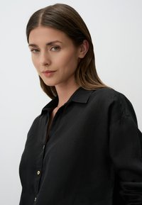 Camicia in lino nera con colletto, caratterizzata da una chiusura con bottoni e vestibilità morbida. La texture appare morbida, con un leggero lucido e dettagli minimi.