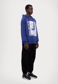 Μπλε φούτερ με γραφικό σχέδιο μπροστά και υφή, σε συνδυασμό με μαύρες φόρμες με μοτίβο και μαύρα αθλητικά παπούτσια.