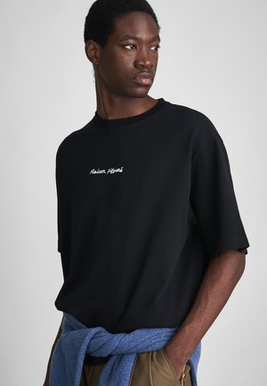 Schwarzes Baumwoll-T-Shirt mit kurzen Ärmeln, das auf der Brust den weißen gestickten Schriftzug "Maison" trägt. Ein gestrickter blauer Pullover ist um die Taille gebunden.