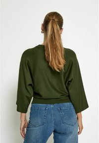 Pull vert à coupe décontractée avec des manches 3/4, associé à un jean bleu taille haute avec des poches standards et une texture en denim.