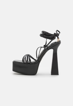 Sandale plateforme noire à talons hauts avec design à lanières, attache à la cheville et talon bloc épais sur fond blanc.