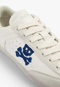 Baskets blanches avec lacets crème, logo tête de mort et os croisés bleu sur le côté, et semelle texturée.