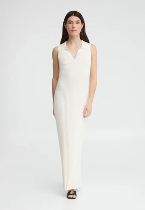 Kobieta o długich brązowych włosach, ubrana w białą, bezrękawną sukienkę maxi z prążkowanego materiału oraz czarne klapki z złotym detalem, stojąca na białym tle.