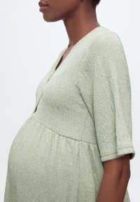 Gravid person i lysegrønn teksturert kjole med V-hals og ermer til midt på armen, vist fra siden mot enkel bakgrunn.