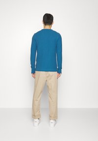 Blå skjorta med långa ärmar och rundad halsringning, kombinerad med cremefärgade chinos och vita sneakers, som visar en avslappnad vy bakifrån.