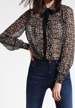 Femme portant un chemisier noir transparent à manches longues avec un motif floral blanc et un détail de nœud noir, associé à un jean bleu foncé taille haute.