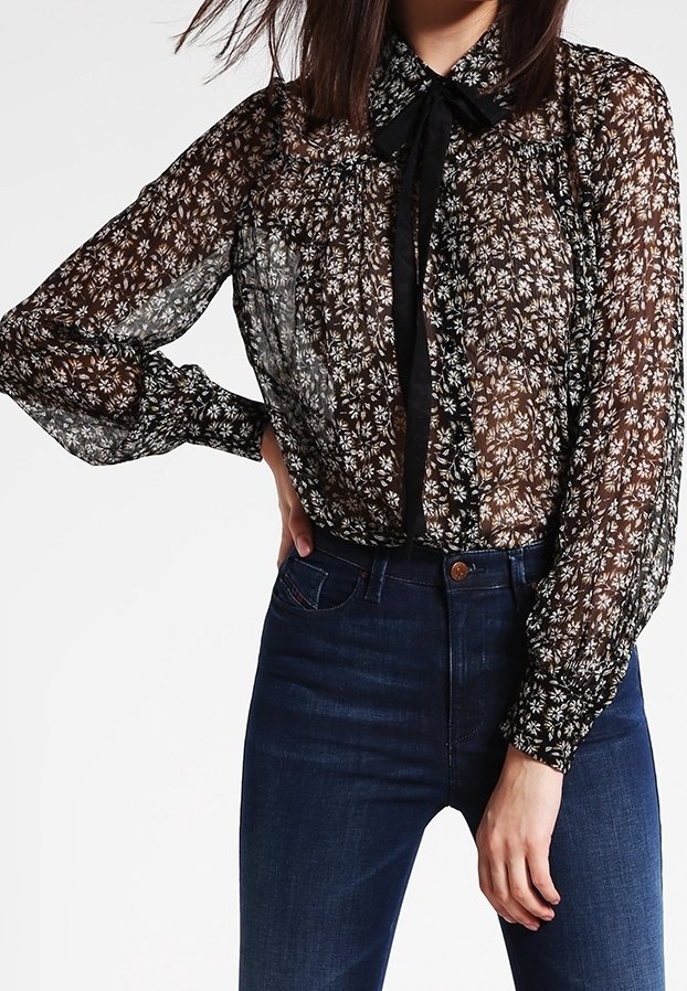 Femme portant un chemisier noir transparent à manches longues avec un motif floral blanc et un détail de nœud noir, associé à un jean bleu foncé taille haute.