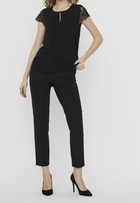 Black top with lace cap sleeves and keyhole detail, paired with black tailored pants. Model wearing black high heels, standing against a white background.