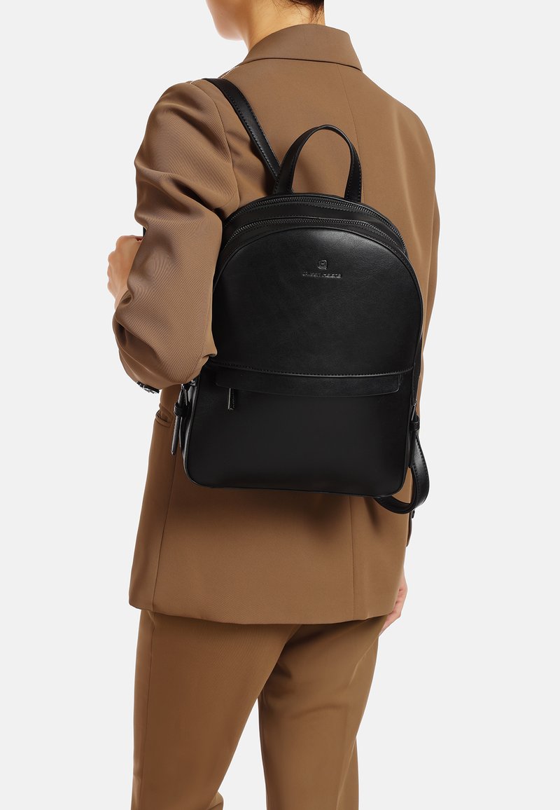 Czarna skórzana plecak o zaokrąglonym kształcie, z kieszonką z przodu, podwójnymi zamkami błyskawicznymi i regulowanymi paskami, noszony na beżowym stroju.