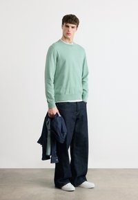 Jeune homme portant un pull vert menthe, un jean large foncé, des baskets blanches, tenant une veste en denim foncé sur un fond blanc uni.