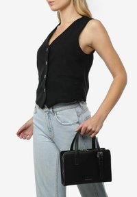 Sort læder håndtaske med dobbelte håndtag og lynlås. Har en glat tekstur og diskret branding på fronten. Båret med en sort vest og lyse jeans.